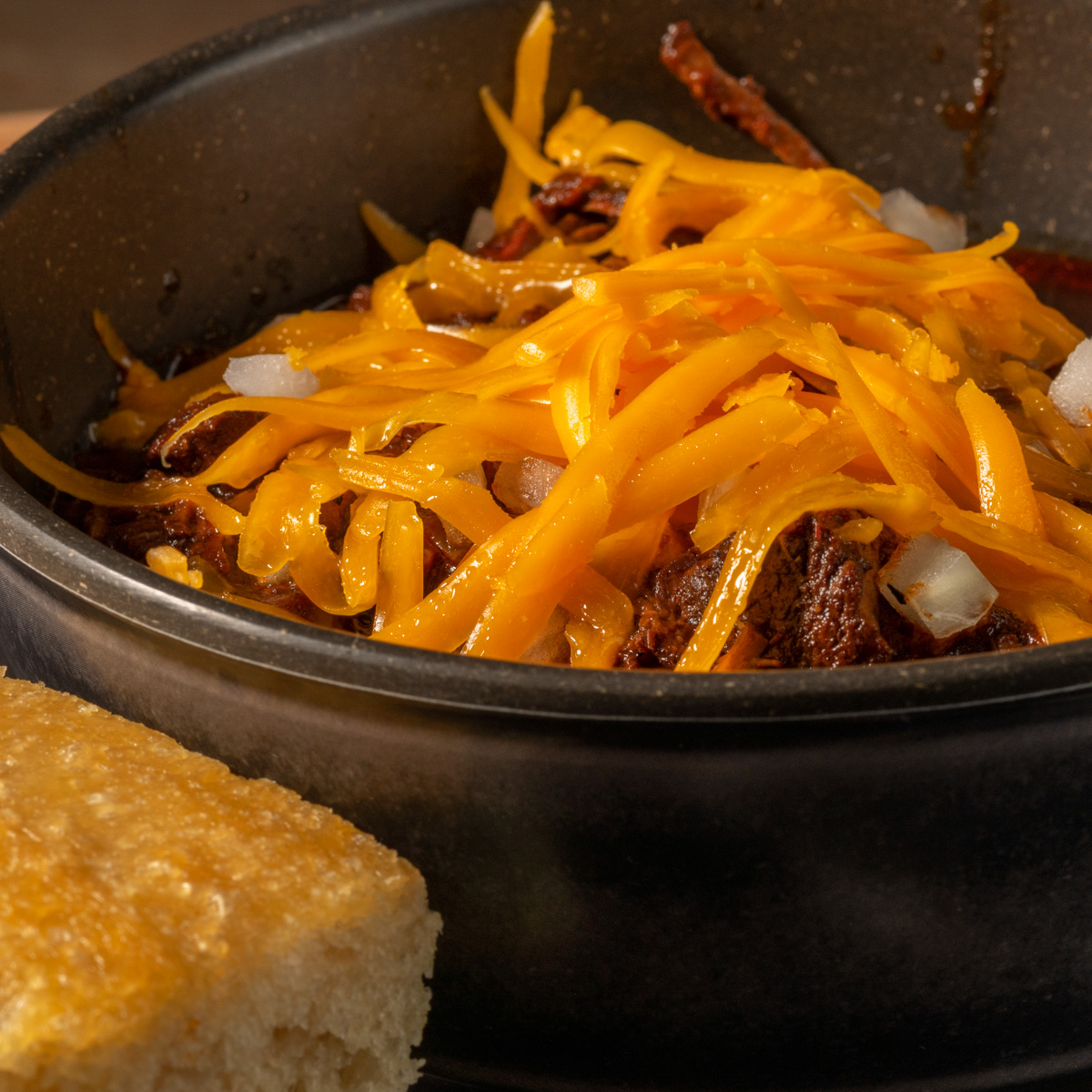 A big bowl of Texas chili topped with shredded cheddar cheese and served with cornbread.