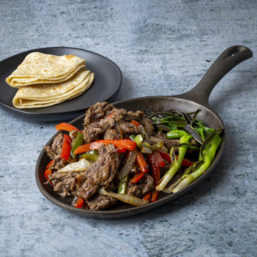 Steak fajitas on a cast-iron comal with a side of warm flour tortillas.